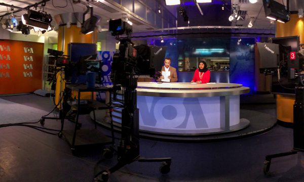 Two VOA television broadcasters sit at a news desk in a studio in front of several cameras