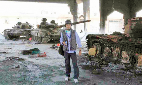 A reporter in tactical gear stands next to a burned out tank with two others in the background