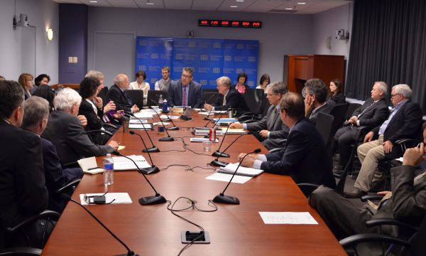 Several men sitting around a conference table with microphones. Several others sit behind them.