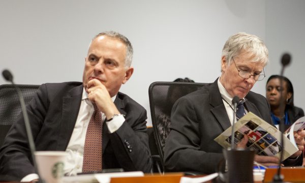 Two men in suits sit at a conference table in front of microphones while the one on the right looks through a book.