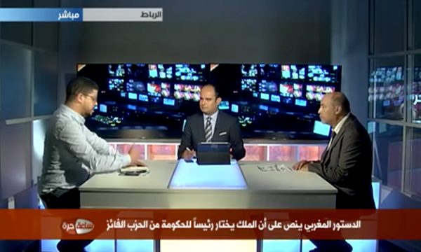 Three men sit at a desk in a television studio during a broadcast