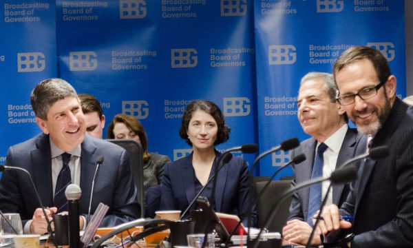 Several men in suits sit at a table in front of microphones during a meeting.