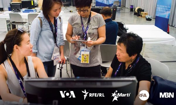 Four Asian women sit or stand in front of a computer screen while having a discussion