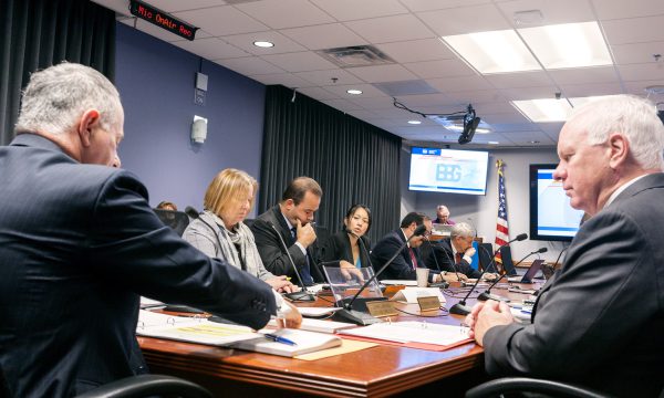 A man sits at the head of a conference table, five men and women sit to his left and a man to his right.