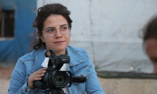 young woman in denim jacket wearing glasses behind a tripod and camera