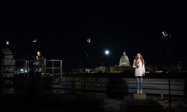 Woman standing on the rooftop of a building holding a microphone, overlooking the U.S. Capitol.